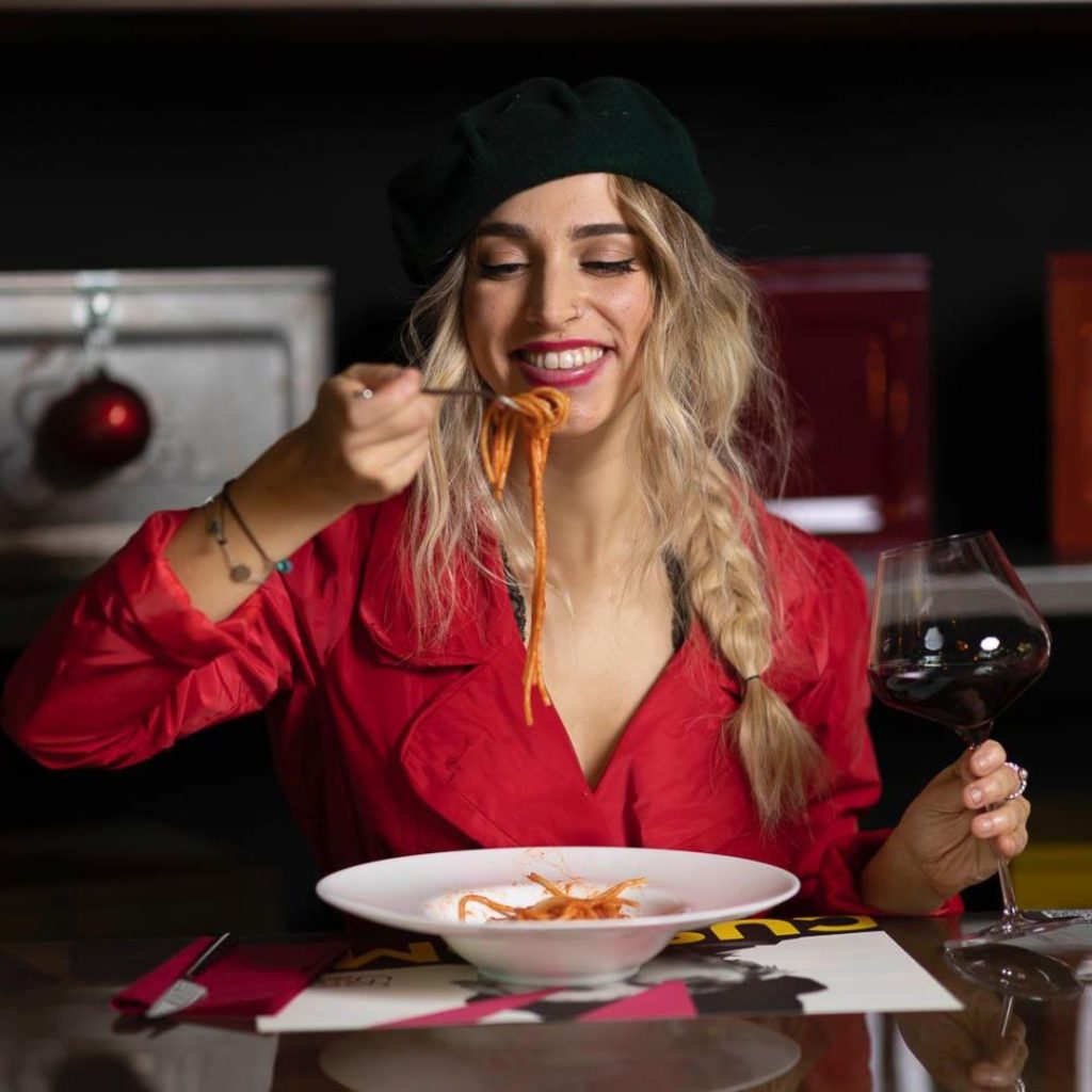 Primo piano di Giorgia Bonafini seduta a tavola intenta a mangiare un piatto di spaghetti e con un calice di vino rosso in mano