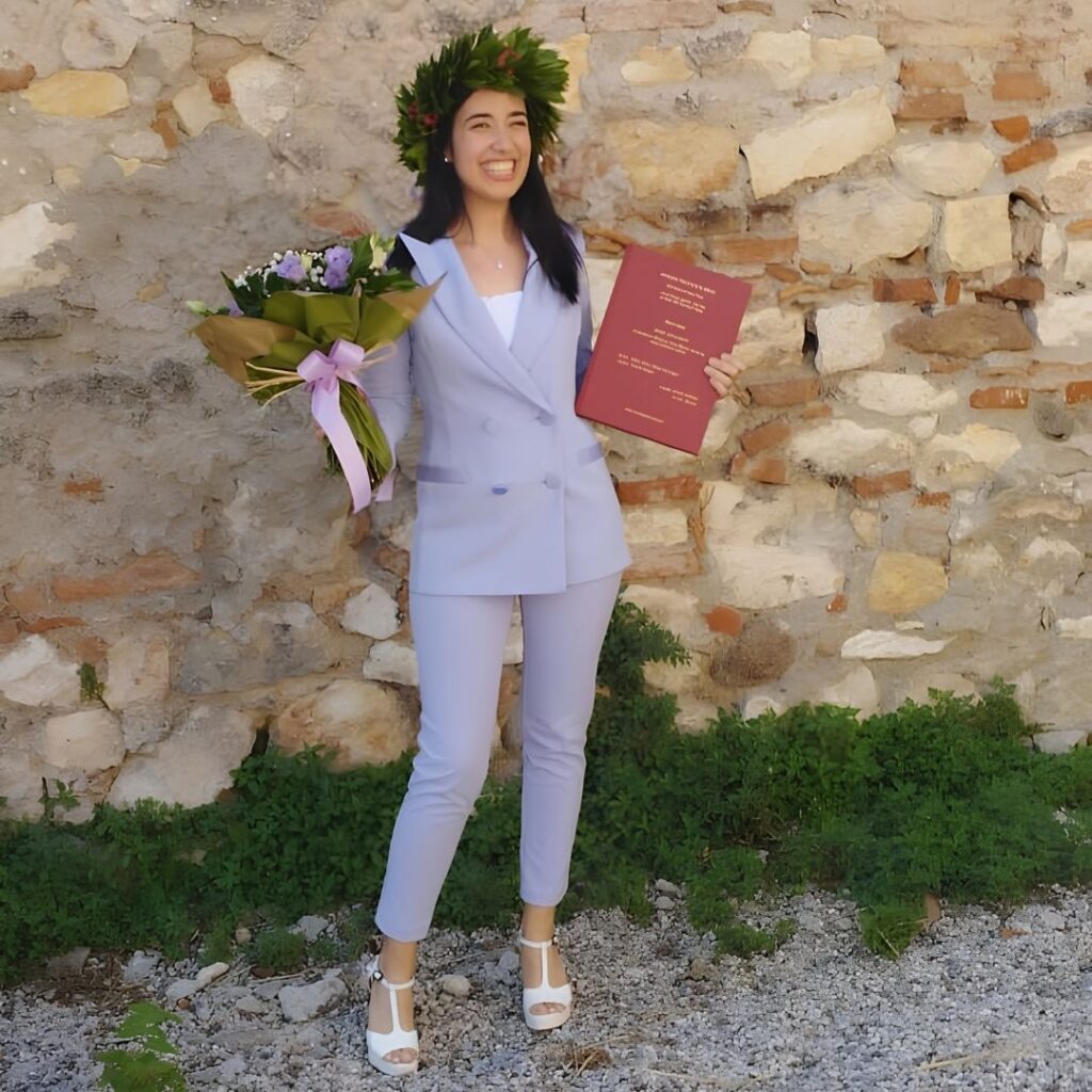 Camilla Pirrello posa in piedi con corona d'alloro e portando tra le mani un mazzo di fiori e la tesi di laurea per festeggiare il conseguimento del titolo universitario